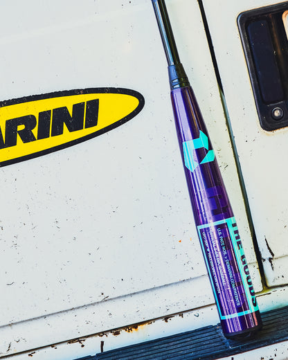 A 2026 DeMarini The Goods Royal Purple (-3) BBCOR Baseball Bat (WBD2634010) by DeMarini leans against a white vehicle door, with part of a yellow and black RINI sticker visible.