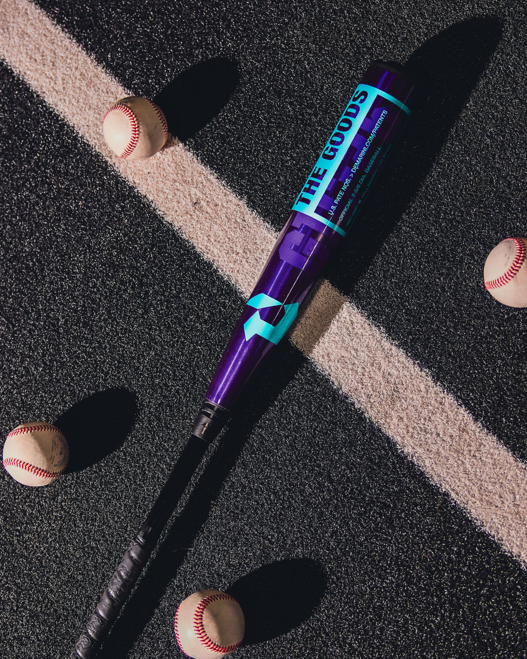 A 2026 DeMarini The Goods Royal Purple (-3) BBCOR Baseball Bat (WBD2634010) from DeMarini rests diagonally on blacktop with white lines, surrounded by five scattered baseballs.