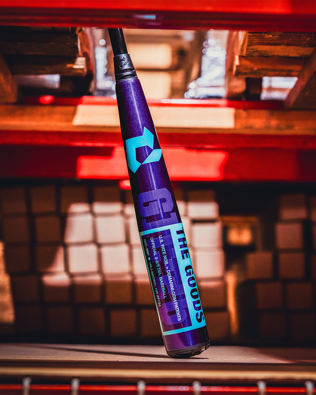 A close-up of the 2026 DeMarini The Goods Royal Purple (-3) BBCOR Baseball Bat (WBD2634010) stands upright against red shelves, with stacked boxes filling the background.