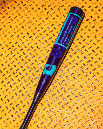 A royal purple 2026 DeMarini The Goods (-3) BBCOR baseball bat (WBD2634010) by DeMarini rests on a yellow textured surface with small square holes, featuring bold blue geometric designs and text.
