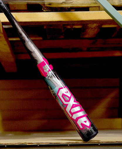 A black and silver DeMarini 2026 Exile Cyborg (-3) BBCOR Baseball Bat (WBD2654010) with bold pink and white "Exile" text rests diagonally against wooden shelves, showcasing its powerful X14 Alloy Barrel in an indoor setting.