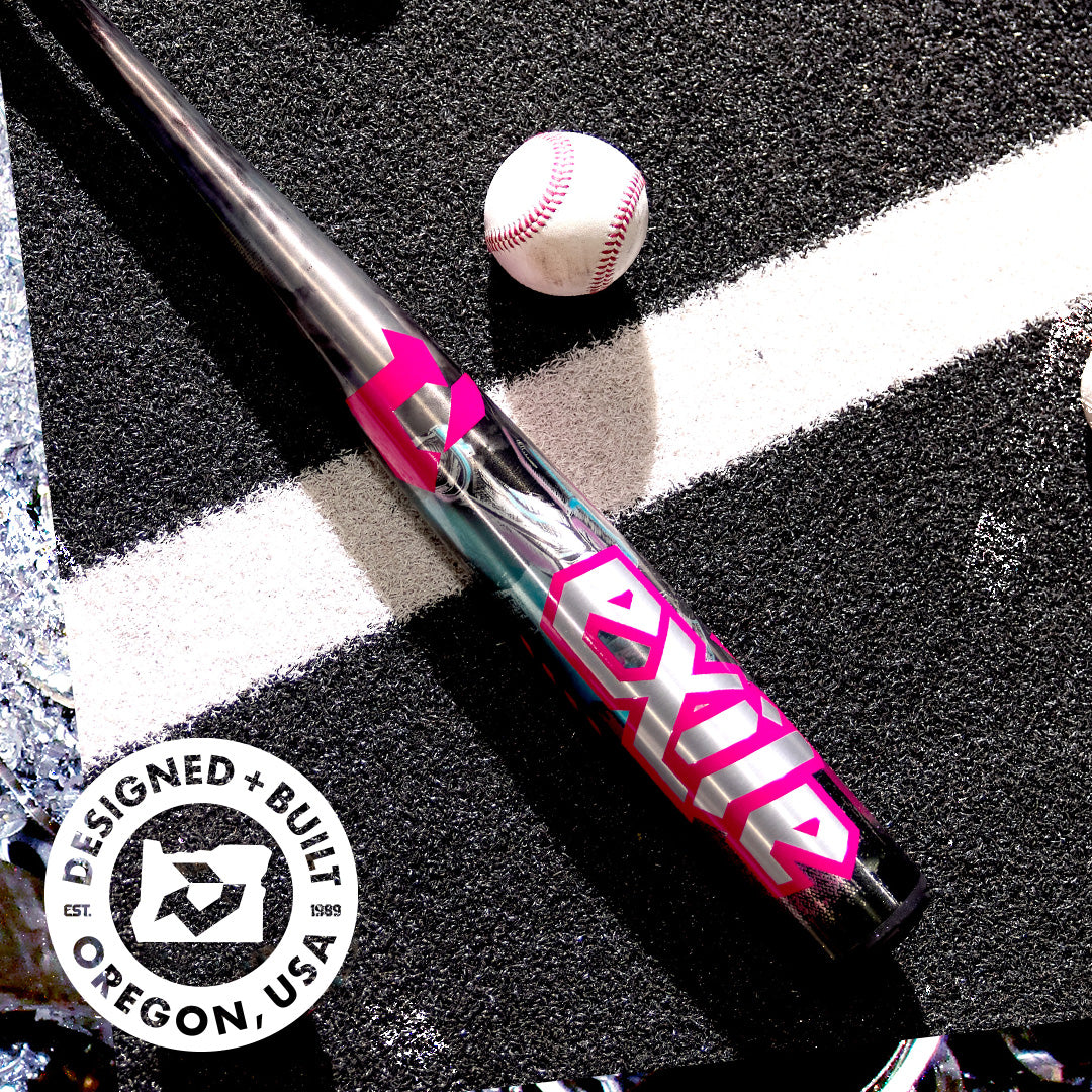 A metallic DeMarini 2026 Exile Cyborg (-3) BBCOR Baseball Bat (WBD2654010), with bold pink and white EXILE letters, lies on asphalt by a white baseball. A circular logo reads DESIGNED + BUILT OREGON, USA in the bottom left corner.