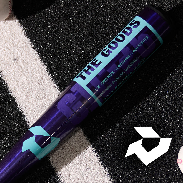 A close-up of a dark purple baseball bat labeled THE GOODS lying on artificial turf near white lines, with a few baseballs and a white geometric logo nearby.
