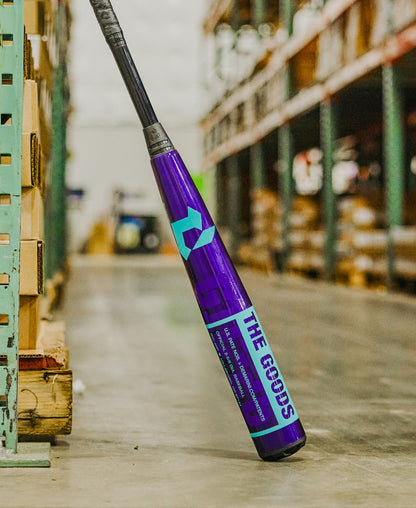 A 2026 DeMarini The Goods Royal Purple (-3) BBCOR Baseball Bat (WBD2634010) from DeMarini stands upright against warehouse shelves, surrounded by stacked boxes and various other stored items.