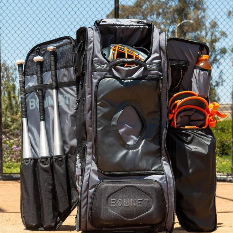 A large open baseball equipment bag with visible compartments holding bats, gloves, helmets, and water bottles, standing on a dirt field with a chain-link fence and trees in the background.