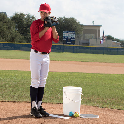 A young baseball player in a red shirt and cap stands on the mound, preparing to pitch. Beside him on the grass is a Champion DSG 6 Gallon Ball Bucket with Padded Lid (BUC6-DSG) by Champion. A scoreboard and American flags are in the background.