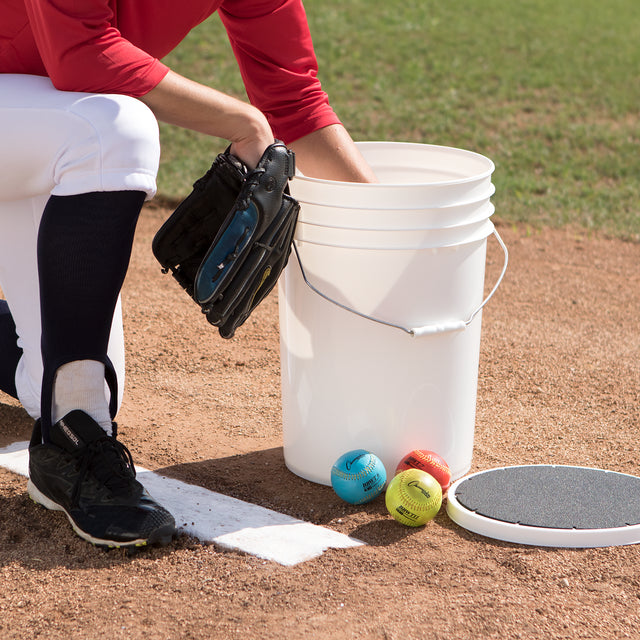 A baseball player in red and white kneels on a dirt field, reaching into a Champion DSG 6 Gallon Ball Bucket with Padded Lid by Champion. Three colorful training balls are nearby, next to a pitching rubber and circular pad.