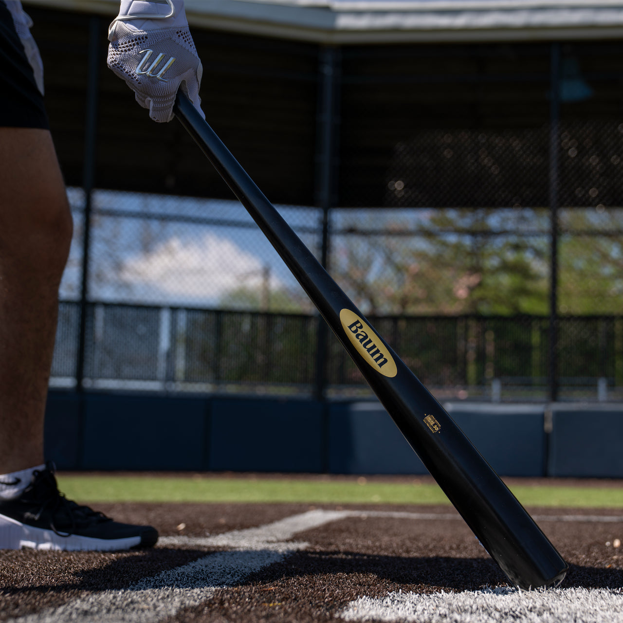 Clad in athletic gear, a person stands near home plate on a baseball field, gripping the Baum Gold Stock AAA Pro Balanced Composite Wood Bat: BBM by Baum, poised for elite performance with a chain-link fence and dugout in the background.