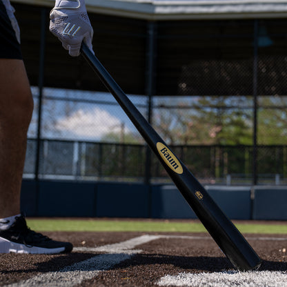 Clad in athletic gear, a person stands near home plate on a baseball field, gripping the Baum Gold Stock AAA Pro Balanced Composite Wood Bat: BBM by Baum, poised for elite performance with a chain-link fence and dugout in the background.