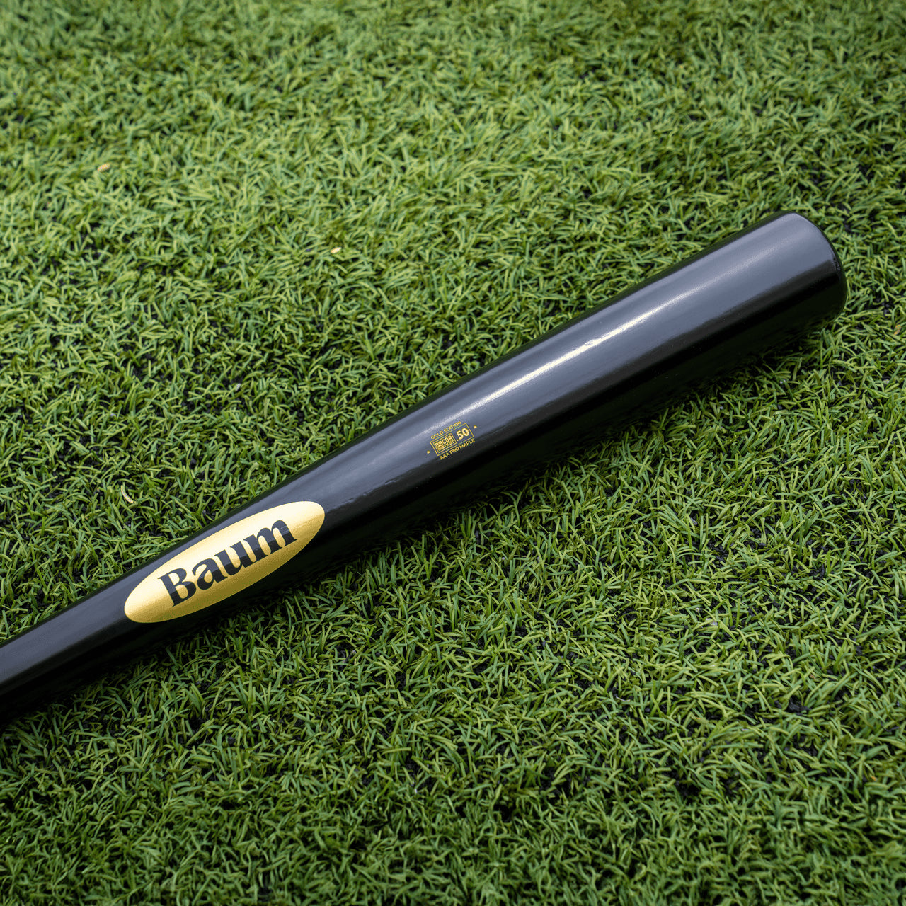 A Baum Gold Stock AAA Pro Balanced Composite Wood Bat: BBM, featuring the iconic yellow Baum logo and expertly handcrafted in the USA for elite performance, rests on bright green artificial turf.