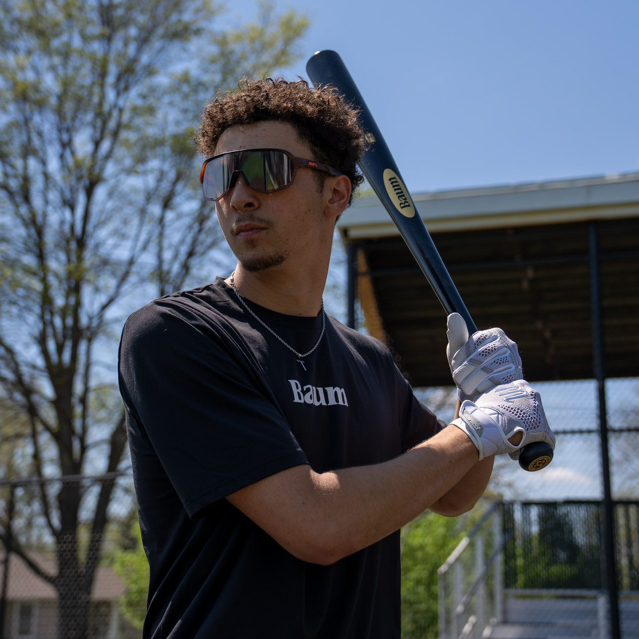 A young man wearing sunglasses and gloves grips a Baum Gold Stock AAA Pro Balanced Composite Wood Bat by Baum, poised to swing on a sunny day near a baseball field surrounded by trees and a fence.
