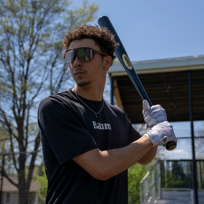 A young man wearing sunglasses and gloves grips a Baum Gold Stock AAA Pro Balanced Composite Wood Bat by Baum, poised to swing on a sunny day near a baseball field surrounded by trees and a fence.