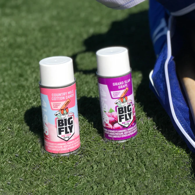 Two cans of All Star Big Fly Bat Tack Spray (one pink Country Mile Cotton Candy, one purple Grand Slam Grape) rest on green artificial grass beside a blue and white All Star bat grip bag.