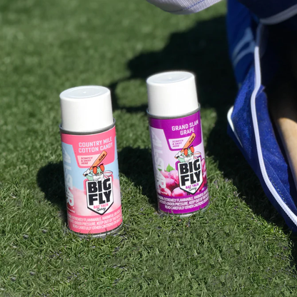 Two cans of All Star Big Fly Bat Tack Spray (one pink Country Mile Cotton Candy, one purple Grand Slam Grape) rest on green artificial grass beside a blue and white All Star bat grip bag.