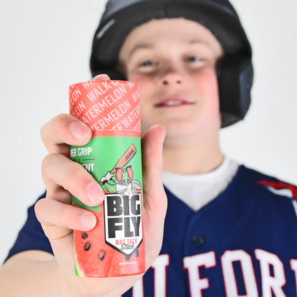A young All-Star in a uniform and helmet holds the All Star Big Fly Bat Tack Stick: BFST1, featuring a watermelon design and showing the product label clearly. The scene has a plain white background.