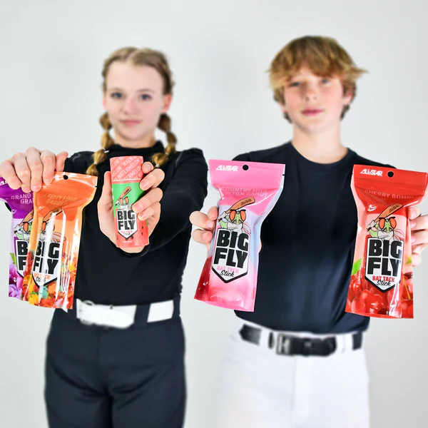 Two kids in black and white uniforms hold up colorful All Star Big Fly Bat Tack Stick (BFST1) packages toward the camera. Against a plain white background, the vibrant packaging features fruit graphics, bold logos, and All-Star bat grip-inspired designs.