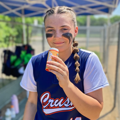A softball player in a blue and white uniform smiles with eye black on, holding a sports drink and displaying her All Star Big Fly Bat Tack Stick (BFST1) under a canopy at an outdoor field on a sunny day.