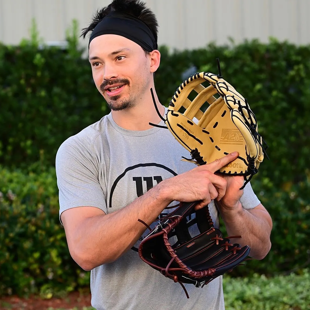 A man wearing a gray t-shirt and black headband holds two baseball gloves, one tan and one brown, while standing outdoors and pointing at the tan glove, with greenery in the background.