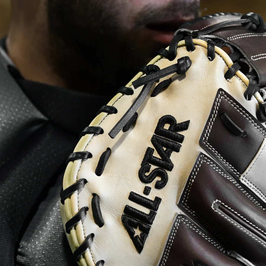 A close-up shows a person in a dark uniform holding the All Star S7 Elite 34" Baseball Catcher's Mitt: CM5000, with "ALL-STAR" embroidered in black and the lower part of their face visible behind the premium-grade rawhide glove.