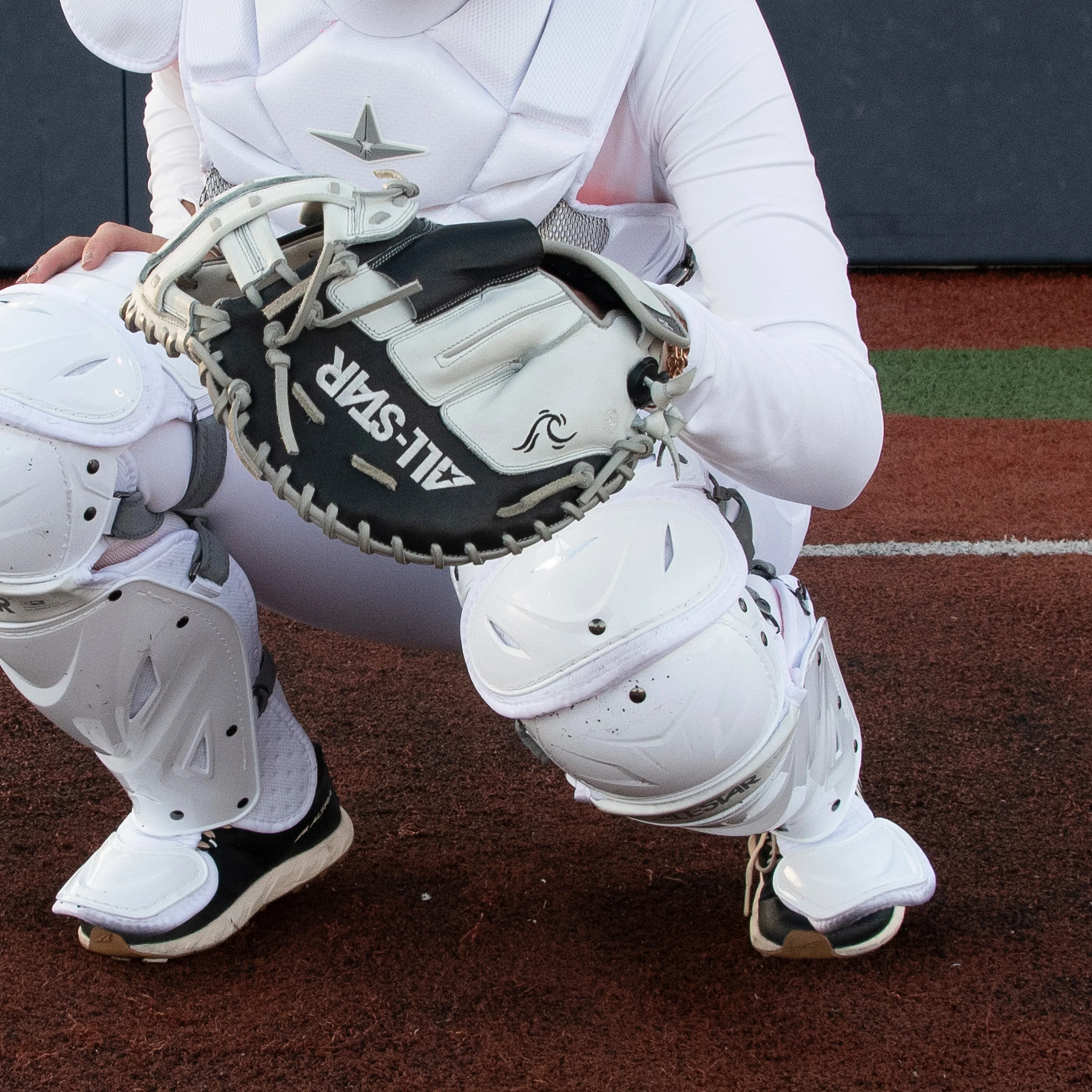 A baseball catcher in white protective gear crouches on a field, holding an open black-and-white mitt marked ALL-STAR, ready to catch a pitch. The background shows the edge between artificial turf and dirt.