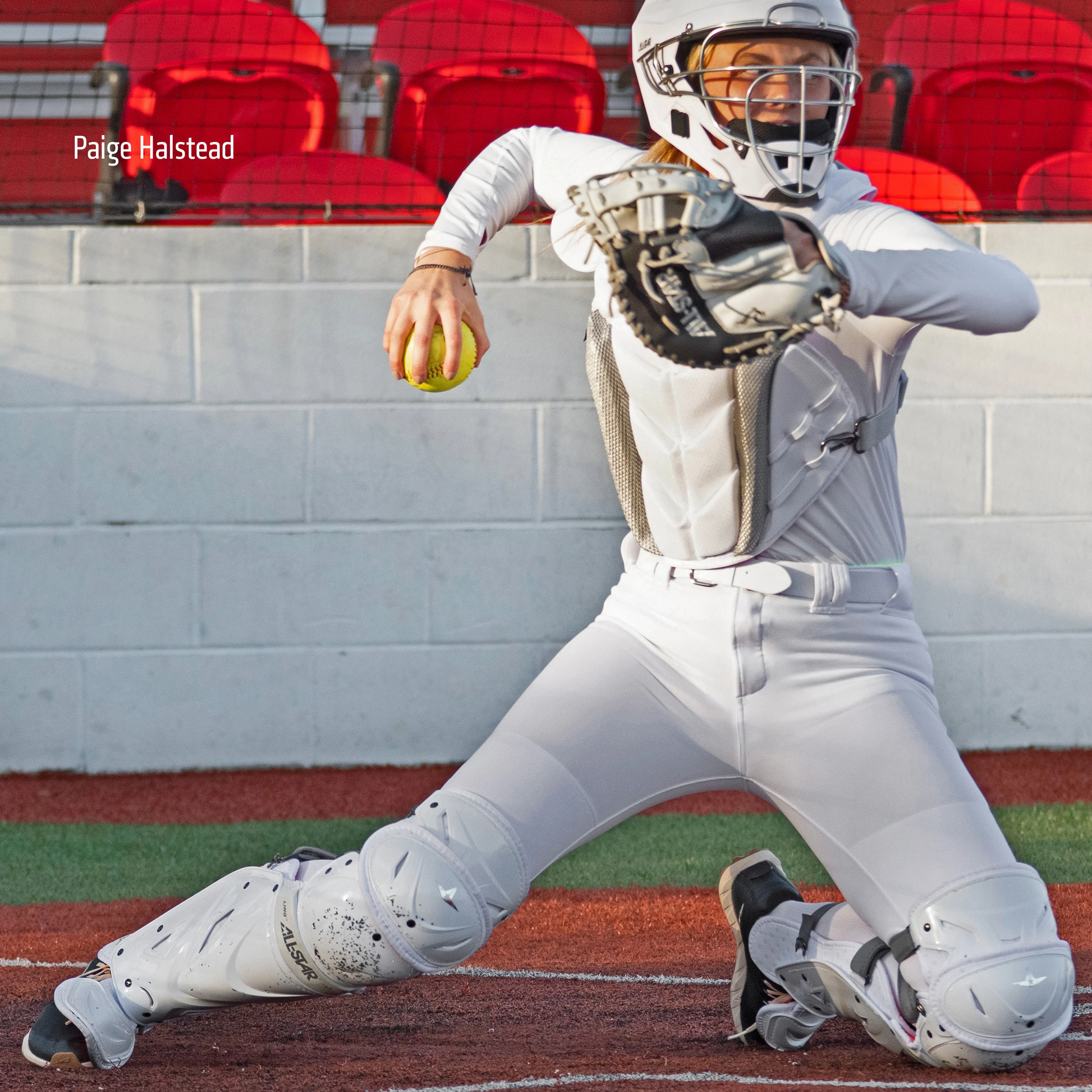 A catcher in All Star PHX Paige Halstead Fastpitch Catcher's Leg Guards (LGW-PHX) prepares to throw a yellow softball behind home plate, with red stadium seats in the background. The name Paige Halstead appears on the image.