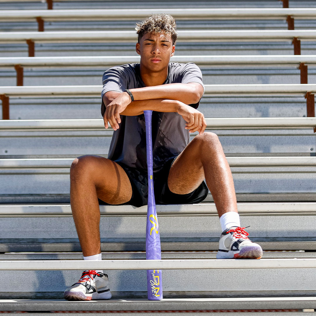 A young man in sportswear sits on stadium bleachers with a Rawlings 2027 Icon The Chosen One (-3) BBCOR Baseball Bat (RBB7IONE3) standing upright between his legs. He rests his arms on the bat and looks at the camera.