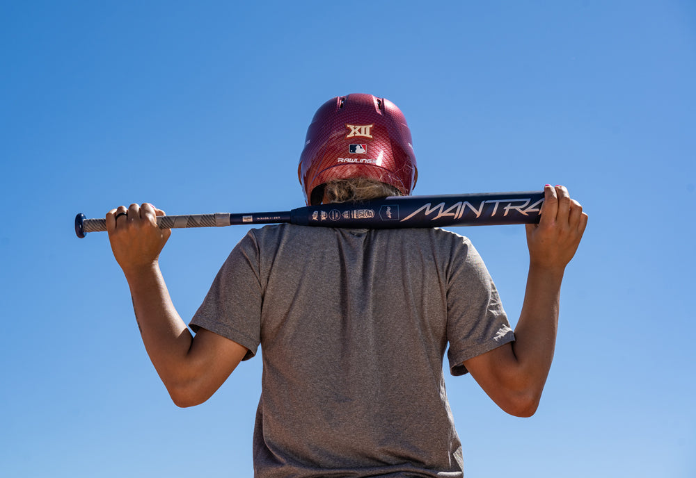 A person in a red helmet and gray shirt holds the 2025 Rawlings Mantra (-9) Fastpitch Softball Bat: RFP4M9 (DEMO) with In/Tense Carbon Composite across their shoulders, facing away under a clear blue sky.