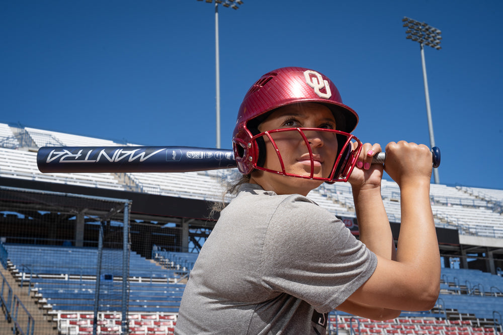 A softball player in a red OU helmet grips a 2025 Rawlings Mantra (-9) Fastpitch Softball Bat (RFP4M9 DEMO), ready to swing on a sunny day at an empty stadium.