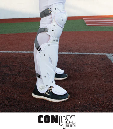 A baseball player wearing All Star PHX Paige Halstead Fastpitch Catcher's Leg Guards (LGW-PHX) with a Delta-Flex calf harness and black athletic shoes stands on a brown and green field. The text "CON4M FIT TECH" appears at the bottom of the image.