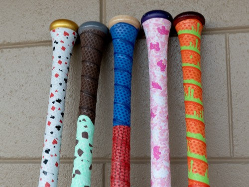 Five baseball bats with colorful, patterned grips resembling ice cream cones and desserts are propped against a tan brick wall. Each bat has a unique design with sprinkles, chocolate, and waffle cone patterns.