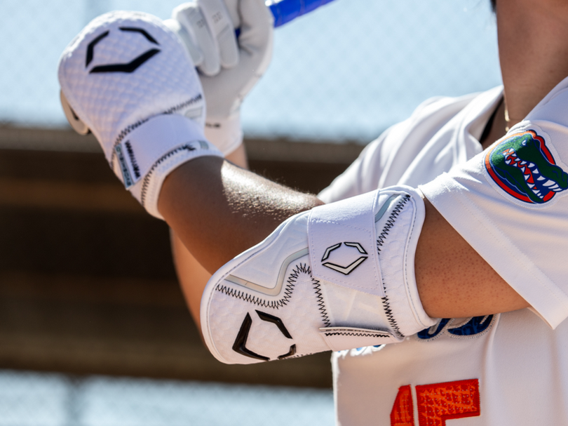 A baseball player wearing white gloves and an elbow guard prepares to bat, holding a blue bat. The players jersey features a patch with an alligator logo on the sleeve.