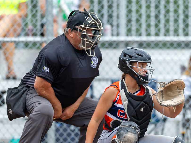 A softball umpire in black gear stands behind a crouched catcher in orange and black uniform, both focused on the field during a game near a chain-link fence.