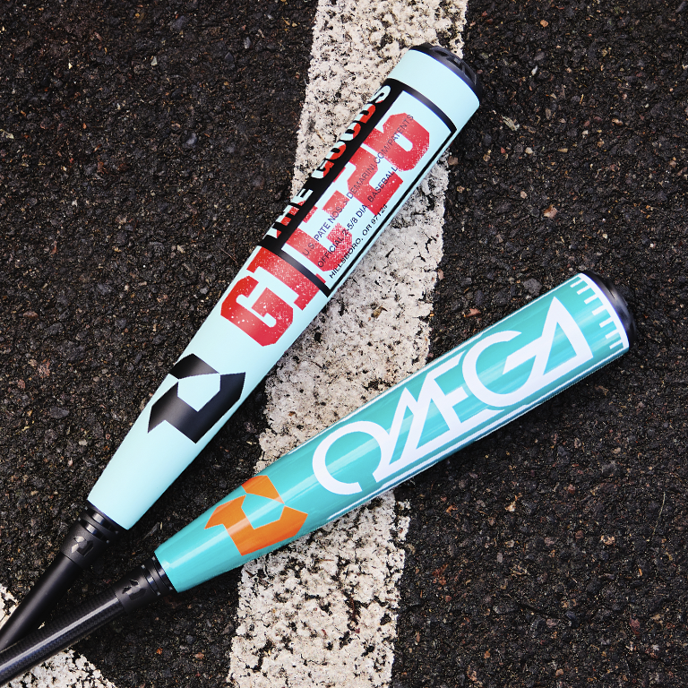 Two blue DeMarini baseball bats—THE GOODS GLITCH and the 2026 DeMarini Omega (-3) BBCOR Baseball Bat: WBD2586010—featuring bold graphics, lie crossed on outdoor asphalt beside a white painted line.