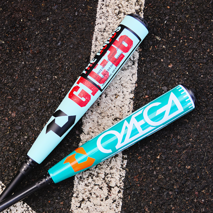 Two blue DeMarini baseball bats—THE GOODS GLITCH and the 2026 DeMarini Omega (-3) BBCOR Baseball Bat: WBD2586010—featuring bold graphics, lie crossed on outdoor asphalt beside a white painted line.