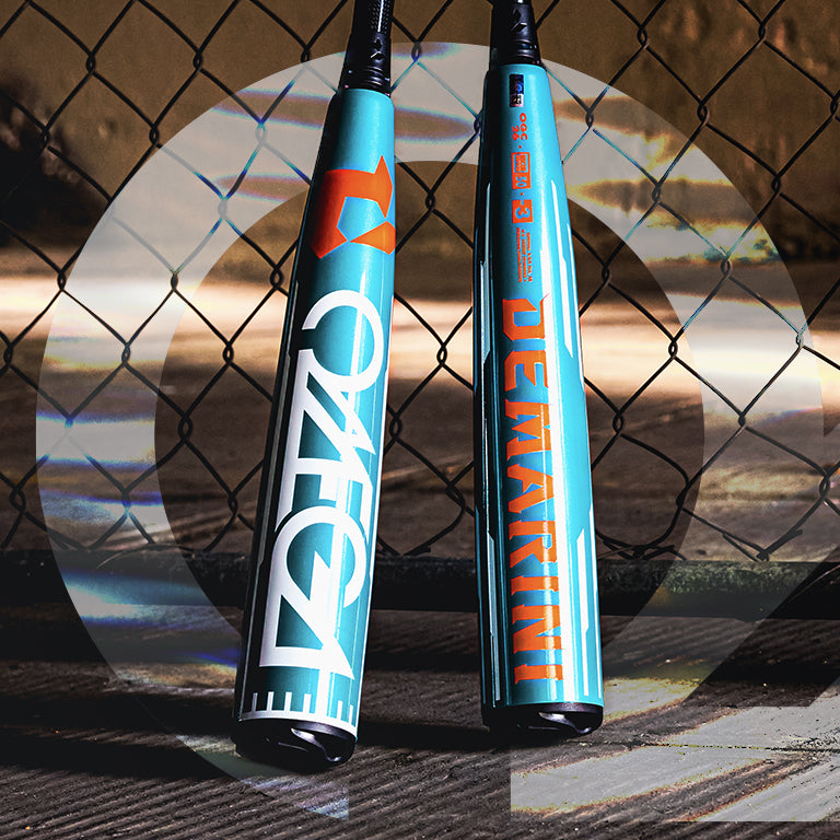 Two teal and black DeMarini baseball bats rest upright against a chain-link fence, with sunlight casting shadows on the ground. Bold “VOODOO ONE” and “DeMARINI” logos are visible on the bat barrels.