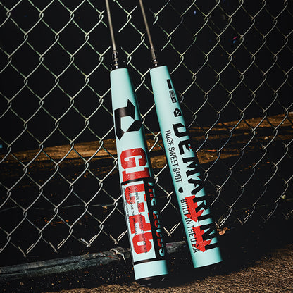 Two 2026 DeMarini The Goods (-3) BBCOR Baseball Bats (WBD2588010) featuring bold red and black graphics lean against a chain-link fence on sunlit concrete, with striking shadows from the fence cast on the ground.