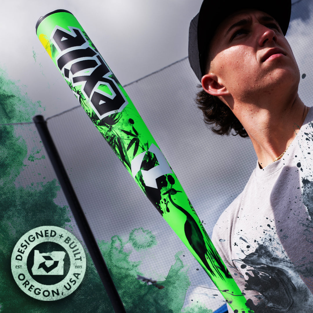 A person in a cap holds the 2026 DeMarini Exile Samurai (-3) BBCOR Baseball Bat: WBD2652010, featuring bold graphics and an X14 Alloy Barrel. A “Designed + Built in Oregon, USA” stamp appears bottom left, set by a fence under a cloudy blue sky.