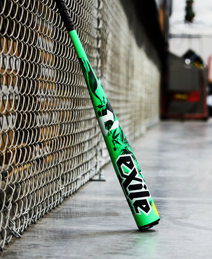 A vibrant green and black 2026 DeMarini Exile Samurai (-3) BBCOR Baseball Bat (WBD2652010) leans against a chain-link fence on concrete, with an industrial backdrop in the distance.