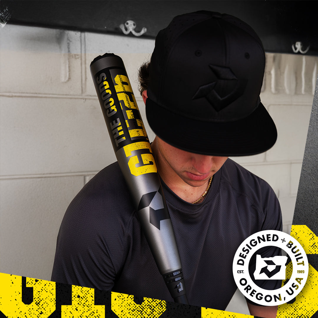 A baseball player in a black cap and shirt rests a DeMarini 2026 The Goods Gunmetal (-3) BBCOR Bat (WBD2650010) on his shoulder, featuring an X14 Alloy Barrel and a circular DESIGNED + BUILT OREGON, USA logo.