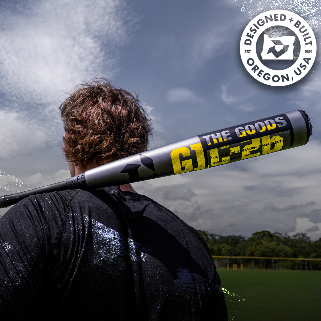 A baseball player stands on a field with the 2026 DeMarini The Goods Gunmetal (-3) BBCOR Baseball Bat (WBD2650010) resting on their shoulder. A circular Designed + Built in Oregon, USA logo appears in the top right corner.