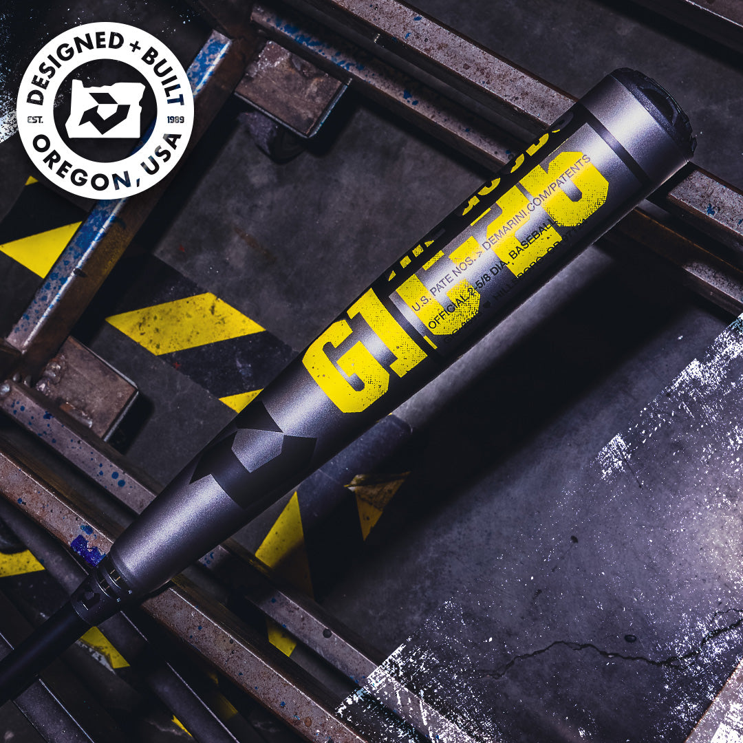 Close-up of a DeMarini 2026 The Goods Gunmetal (-3) BBCOR Baseball Bat (WBD2650010), featuring gray and yellow design with G143 marking; construction materials and caution-striped floor in background. "Designed + Built Oregon, USA" logo shown.