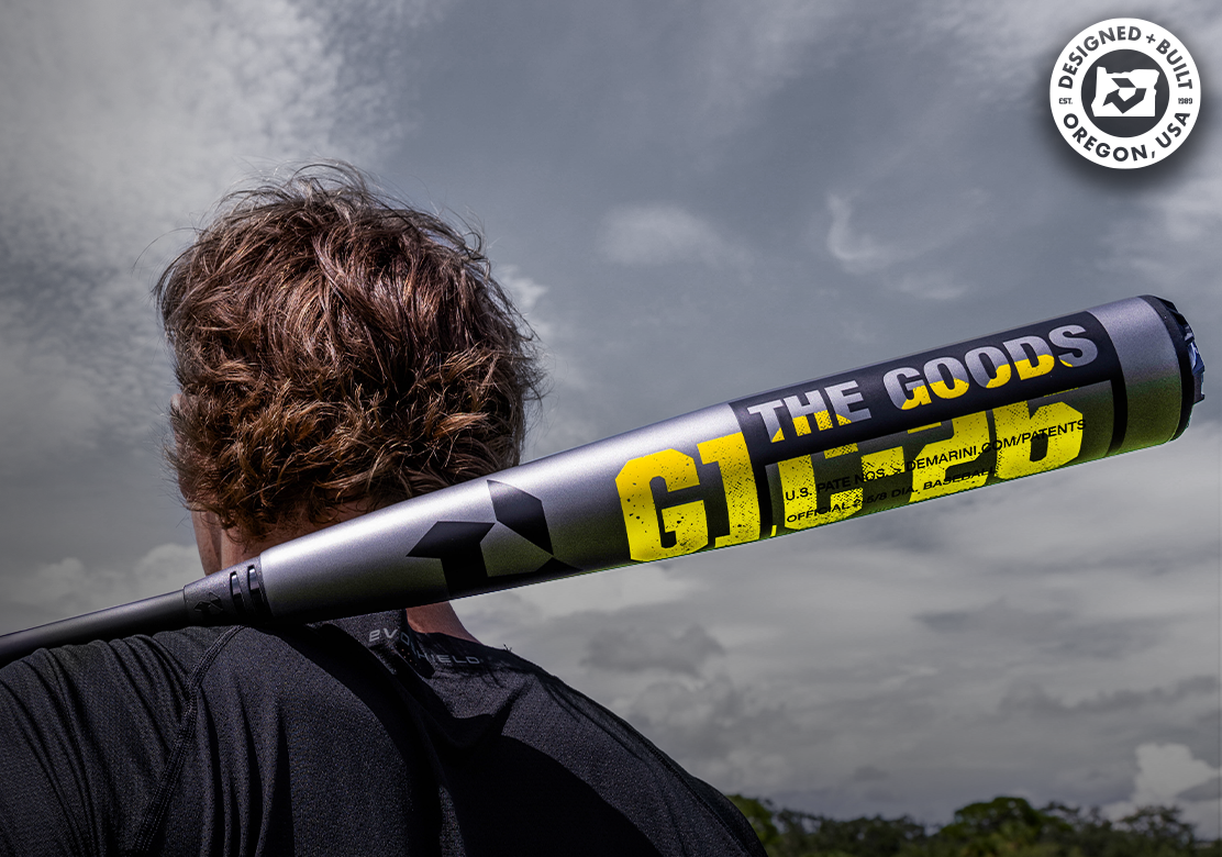 A person with brown hair holds a gray and yellow baseball bat labeled THE GOODS and G1M over their shoulder outdoors. A Designed & Built in Oregon, USA logo appears in the upper right corner.