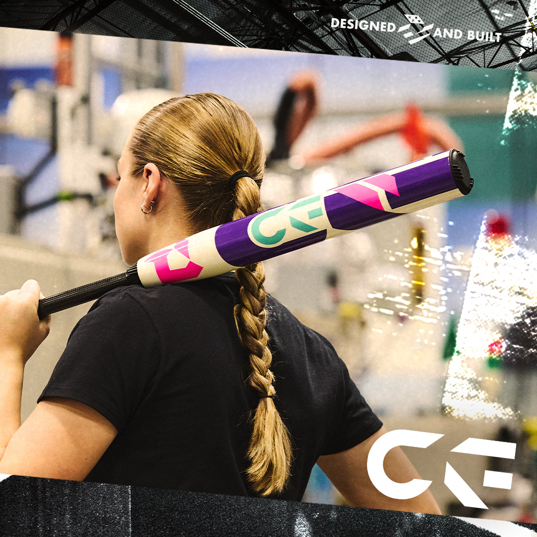 A person with a long braided ponytail holds the 2026 DeMarini CF (-11) Fastpitch Softball Bat: WBD2630010 over their shoulder indoors. The colorful purple bat features pink, green, and white designs, plus a Paraflex Plus Composite Barrel.