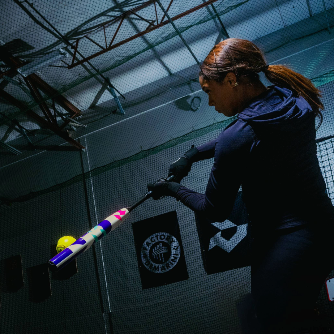 A woman in athletic wear swings a 2026 DeMarini CF (-11) Fastpitch Softball Bat (WBD2630010) at a yellow softball inside an indoor training facility with netting and banners on the walls.