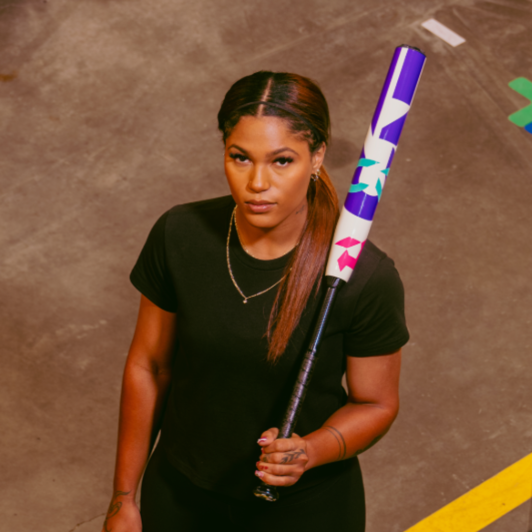 A woman with long, straight hair in a low ponytail, wearing a black t-shirt and gold chain, stands indoors holding the 2026 DeMarini CF (-11) Fastpitch Softball Bat (WBD2630010) over her shoulder, looking up with a serious expression.