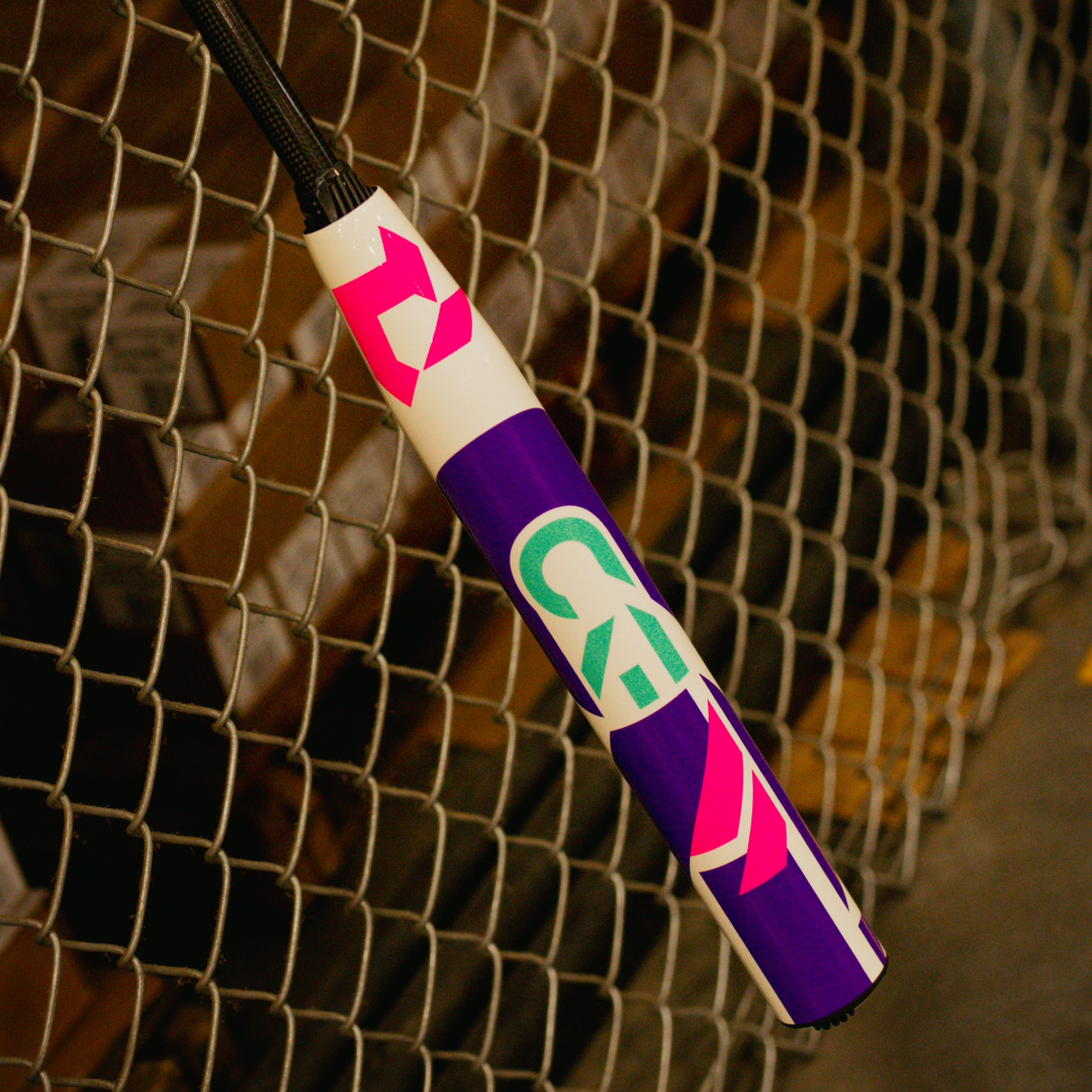 A close-up of the 2026 DeMarini CF (-11) Fastpitch Softball Bat (WBD2630010) displays bold pink, purple, and green graphics and a Paraflex Plus Composite Barrel against a metal chain-link fence, its vivid design standing out.