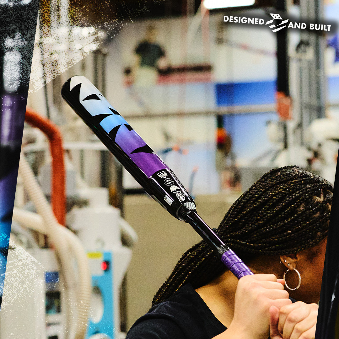 A person with braided hair holds a 2026 DeMarini Prism+ (-11) Fastpitch Softball Bat (WBD2636010) in an indoor workshop. "DESIGNED AND BUILT" appears in the upper right, with machinery and equipment seen in the background.
