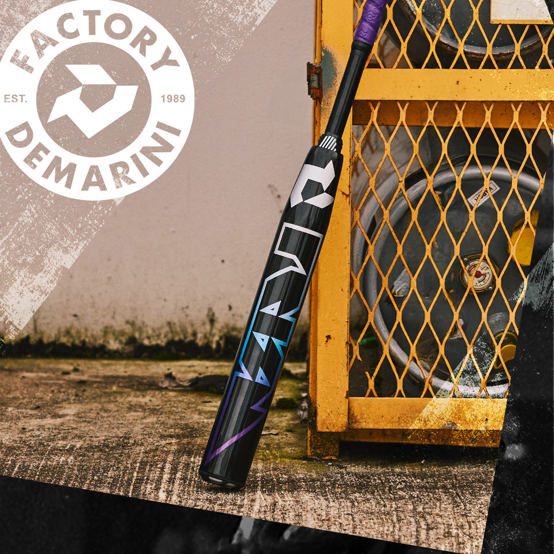 A black 2026 DeMarini Prism+ (-11) Fastpitch Softball Bat (WBD2636010) with colorful accents leans on a yellow metal cage by stacked barrels, displaying the Continuous Fiber Composite Barrel and DeMarini logo.