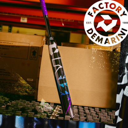 A 2026 DeMarini Prism+ (-11) Fastpitch Softball Bat (WBD2636010) in purple and black stands upright on foam padding in a warehouse, with cardboard boxes and the DeMarini Factory logo in the background.
