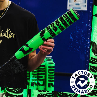 A person in a black shirt holds the neon green and black 2026 DeMarini ZEN (-8) 2 3/4" USSSA Baseball Bat (WBD2592010) with more bats behind them; a circular “Factory DeMarini” logo appears in the bottom right corner.